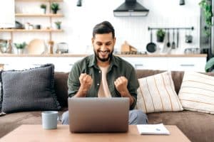 Win, success, triumph. Happy arabian and indian man, sits on a sofa in living room, looks at laptop