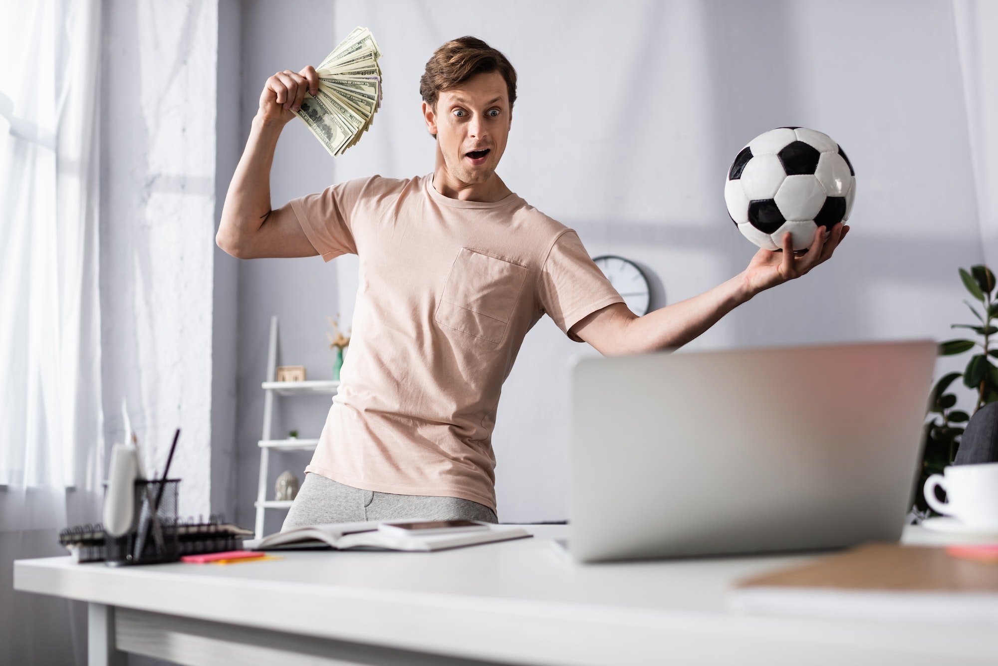 Selective focus of shocked man with cash and football looking at laptop in living room, concept of